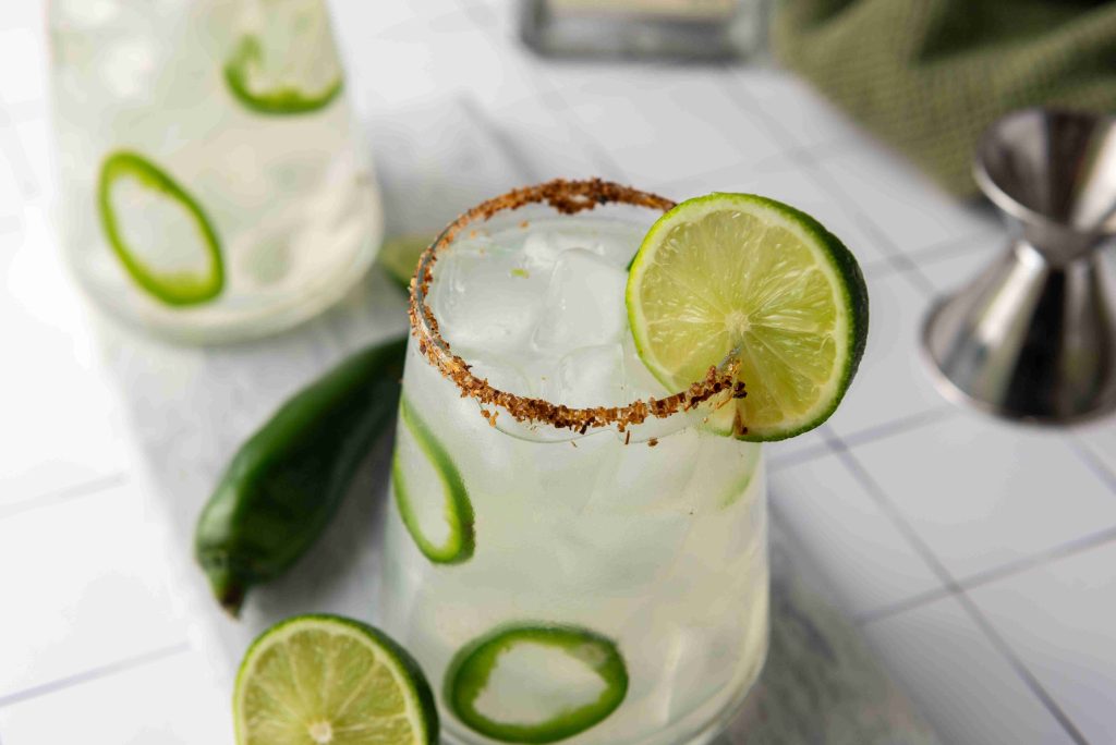 A glass of Spicy Margarita garnished with lime slices and a chili-salt rim, with visible jalapeño slices and Jalapeno Tequila inside; another similar cocktail and fresh jalapeños are in the background on a white surface.