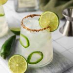A glass of Spicy Margarita cocktail made with jalapeno tequila, garnished with lime slices and jalapeño rings, and a chili-salt rim. Fresh lime and jalapeño sit beside the glass; a metal jigger and green towel are in the background.