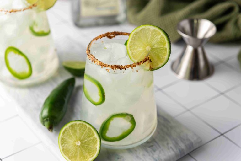 A glass of Spicy Margarita cocktail made with jalapeno tequila, garnished with lime slices and jalapeño rings, and a chili-salt rim. Fresh lime and jalapeño sit beside the glass; a metal jigger and green towel are in the background.