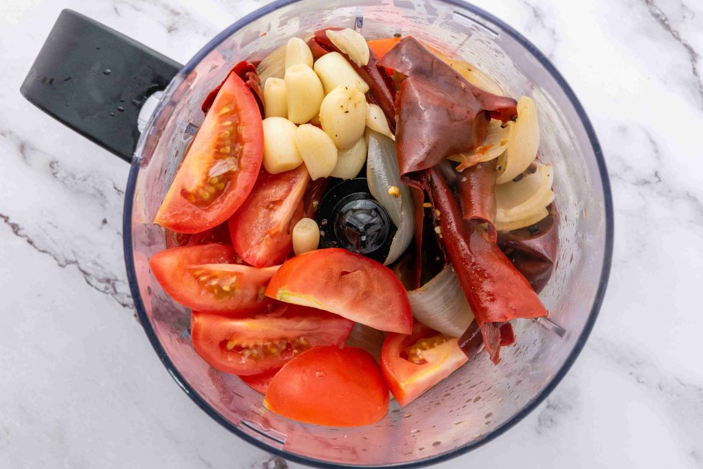 A food processor bowl containing chopped tomatoes, garlic cloves, onion pieces, and dried red chiles for a birria tacos recipe sits ready to be blended on a marble countertop.