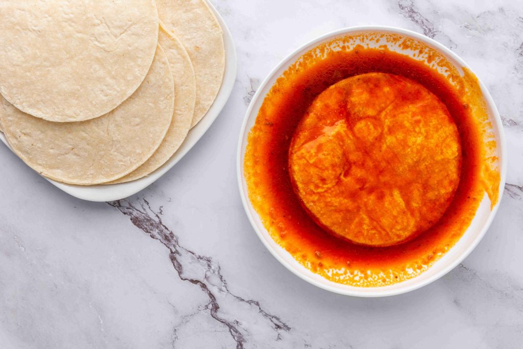 A white bowl filled with red enchilada sauce and a tortilla soaking in it sits on a marble surface next to a plate with a stack of plain corn tortillas, perfect for assembling your favorite birria tacos recipe.