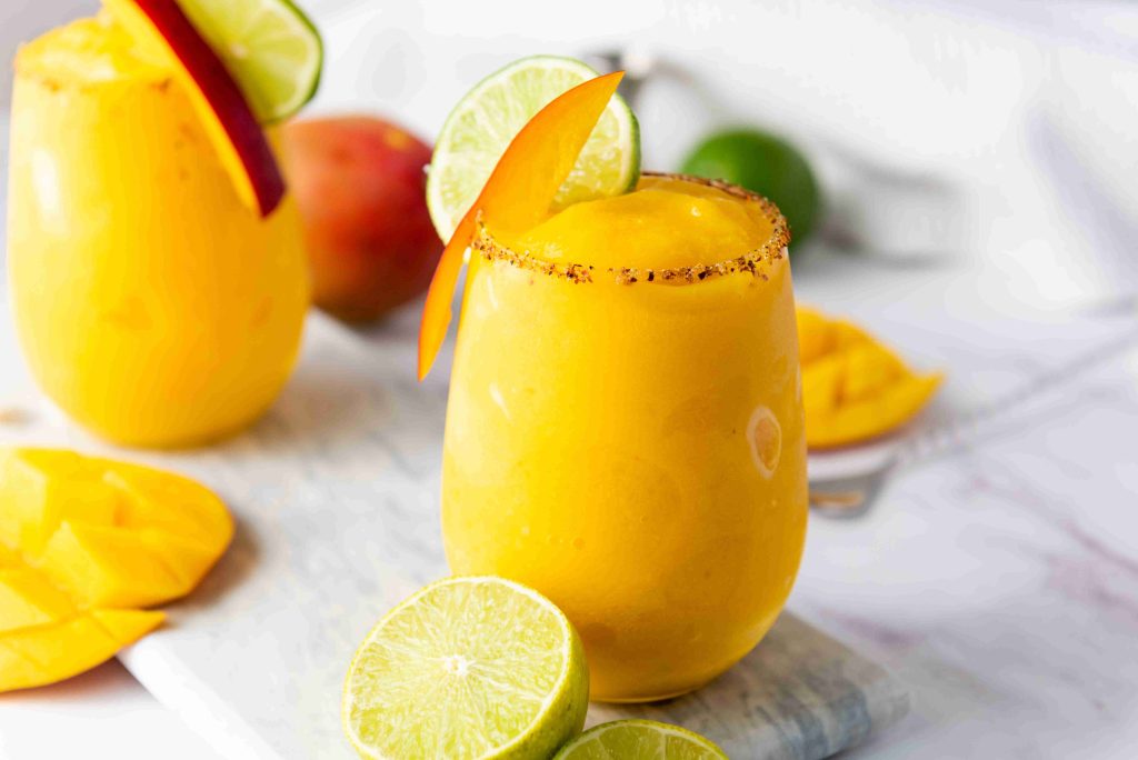 A bright yellow Frozen Mango Margarita in a glass, garnished with a lime slice, mango slice, and chili powder on the rim. Another similar drink, fresh lime, and mango pieces sit in the background on a white surface.