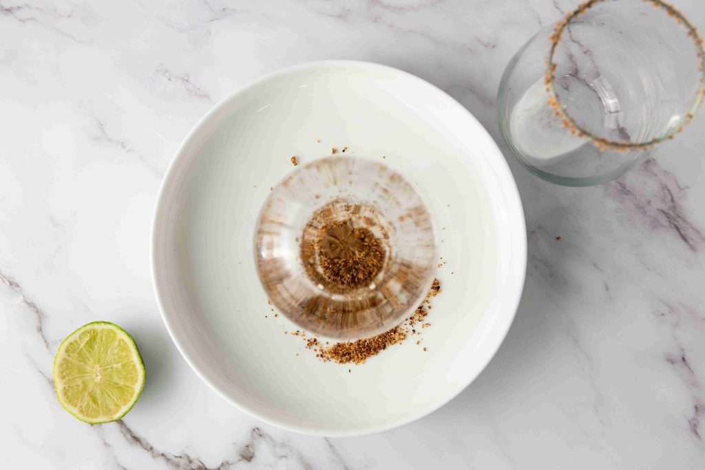 A white plate with brown sugar around an upturned glass, a half-squeezed lime on a marble surface, and an empty glass with a sugared rim beside the plate—perfect prep for a refreshing Frozen Mango Margarita.