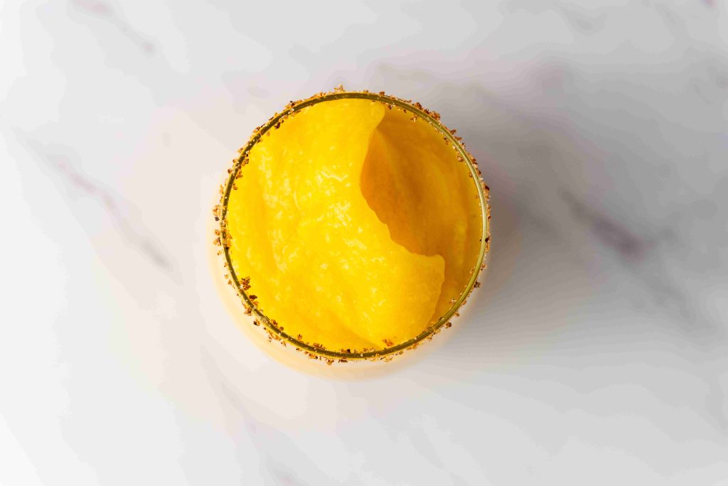A top-down view of a glass filled with bright yellow Frozen Mango Margarita, its spice-coated rim popping against the light marble surface.