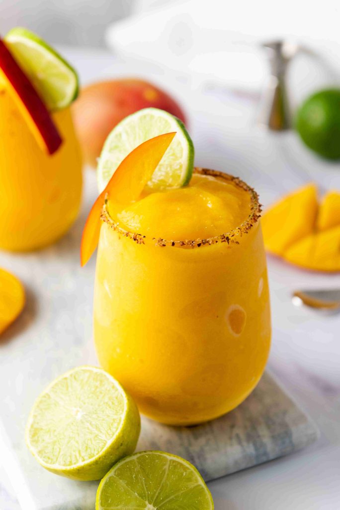 A bright yellow Frozen Mango Margarita in a glass with a chili powder rim, garnished with a lime wheel and mango slice. Fresh lime halves and mango pieces are nearby on a white marble surface.