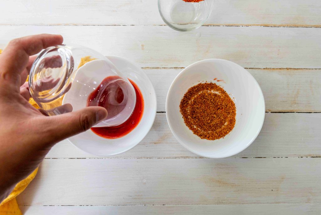 A hand pours a reddish easy mangonada from a glass into a white bowl next to another bowl filled with brown seasoning, all set on a white wooden surface.