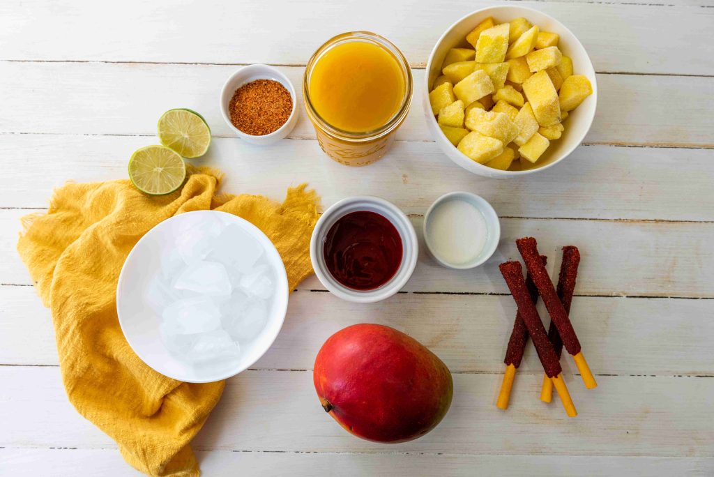 A flat lay of ingredients for an easy mangonada: frozen mango cubes, mango juice, lime, chili powder, ice, honey, milk, tamarind sticks, and a whole mango on a white wooden surface with a yellow cloth. The perfect sweet and spicy drink!.