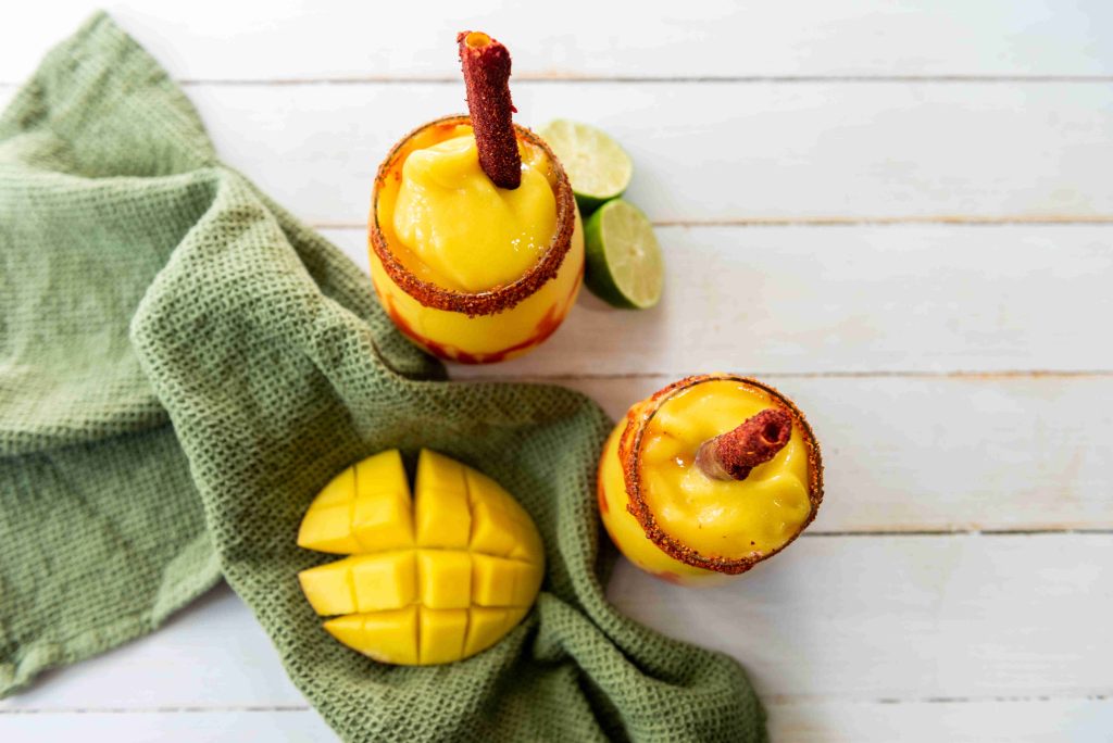 Two glasses of this sweet and spicy drink, inspired by an easy mangonada recipe, sit on a green cloth with a sliced mango and lime halves, their rims dusted with chili powder on a white wooden surface.