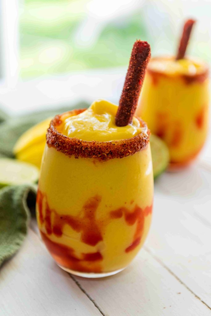 A glass of sweet and spicy drink, inspired by an easy mangonada recipe, with chili powder on the rim, swirls of red sauce inside, and a tamarind candy stick garnish. Another blurred glass sits in the background on a white table.