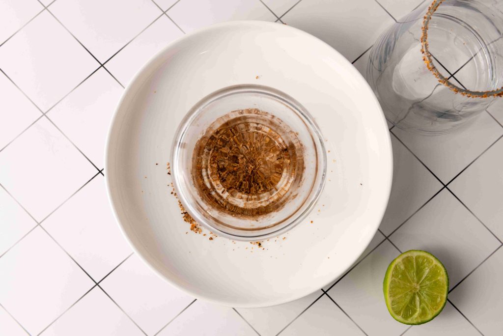 A glass rimmed with spices is placed upside down on a plate, next to a lime half and another glass with a spice-coated rim—perfect for serving a Spicy Margarita or Jalapeno Tequila cocktail—all set on a white tiled surface.
