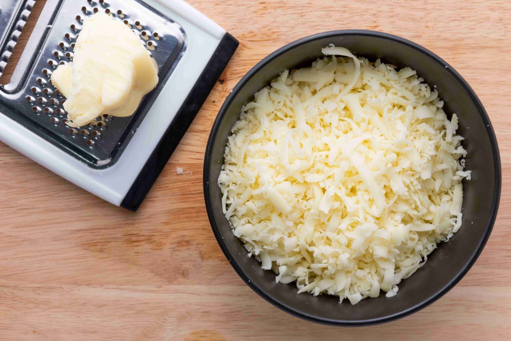 A black bowl filled with shredded cheese sits on a wooden surface next to a metal grater with a block of cheese on top, ready for your next birria tacos recipe.