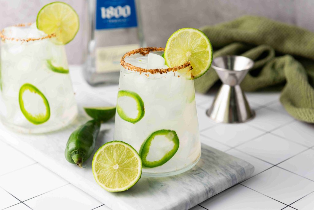 Two glasses of Spicy Margarita, made with jalapeño tequila and garnished with lime and jalapeño slices, sit on a marble board. A bottle of tequila, a jigger, and a green cloth complete this inviting cocktail scene.