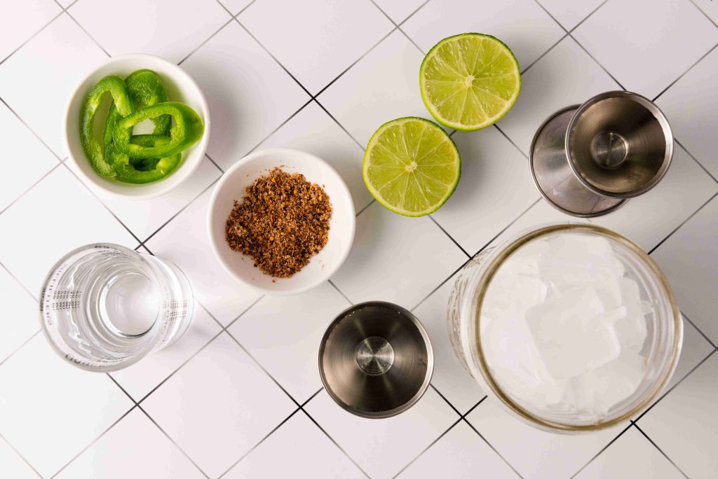 Overhead view of Spicy Margarita cocktail ingredients on a tiled surface: sliced green pepper, lime halves, a bowl of seasoning, Jalapeno Tequila, a glass of water, a jar of ice, and metal measuring jiggers.