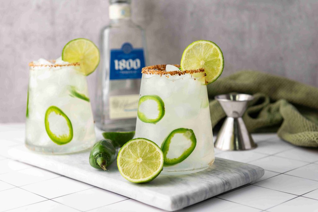Two glasses of Spicy Margarita garnished with lime slices and chili, featuring a bottle of Jalapeno Tequila, a jigger, and a green cloth in the background, all arranged on a marble tray.