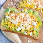 Two slices of whole grain Mexican avocado toast topped with mashed avocado, roasted corn, queso fresco, and a sprinkle of red seasoning, served on a wooden board.