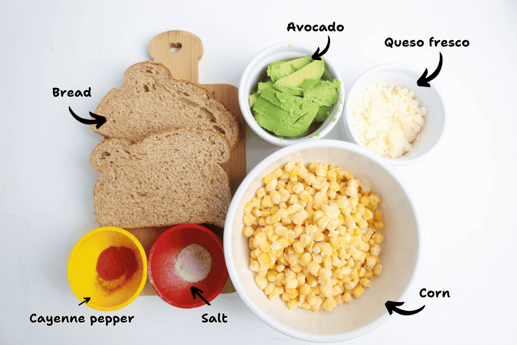 A top-down view of ingredients for Mexican Avocado Toast on a white surface: sliced bread, a bowl of corn, crumbled queso fresco, sliced avocado, and small bowls of cayenne pepper and salt, each labeled.