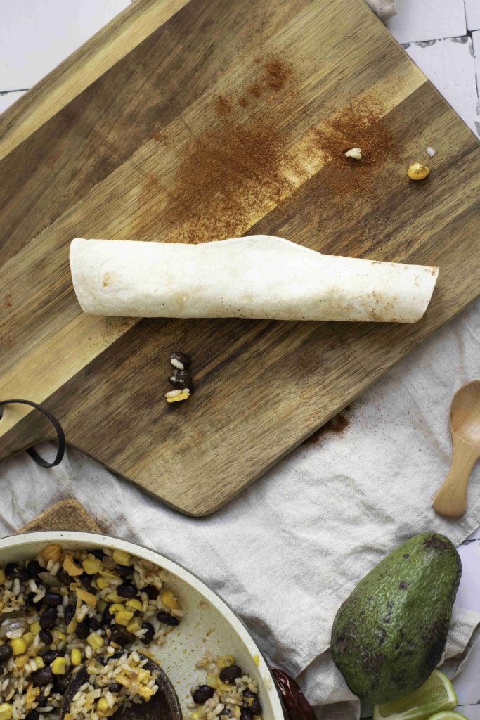 A rolled tortilla sits on a wooden cutting board dusted with spices, next to a pan of beans and rice—perfect for making the best enchiladas. An avocado, lime wedges, and a wooden spoon are nearby on a cloth.