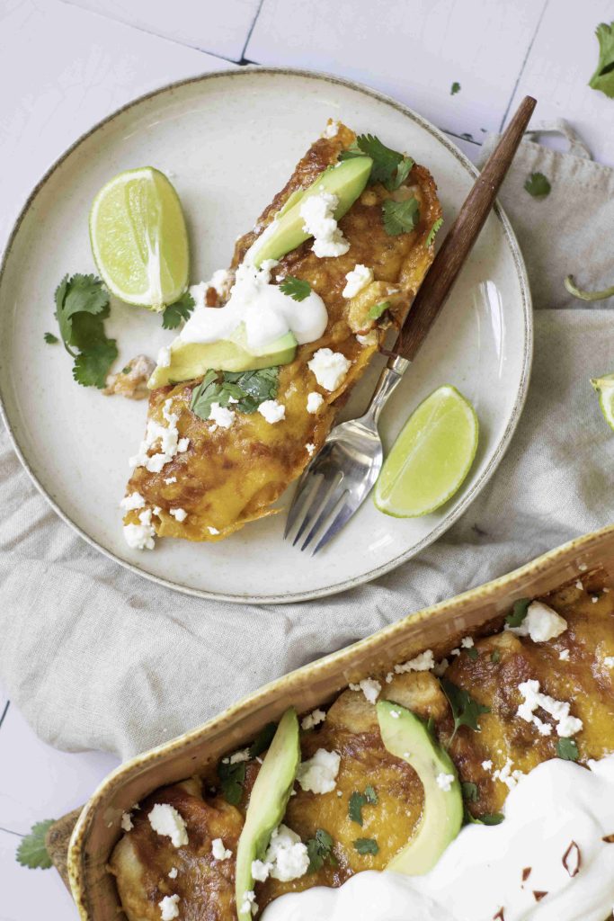 A plate with the best enchiladas topped with avocado slices, cheese, cilantro, and sour cream, served with lime wedges. A casserole dish with more enchiladas is partially visible below. A fork rests on the plate.