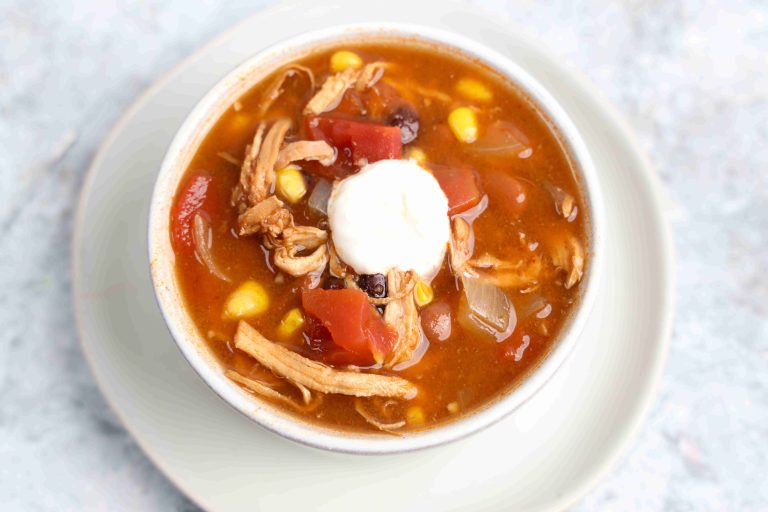 A bowl of Mexican-inspired Chicken Enchilada Soup with shredded chicken, black beans, corn, tomatoes, and onion, topped with a dollop of sour cream, served on a white plate.