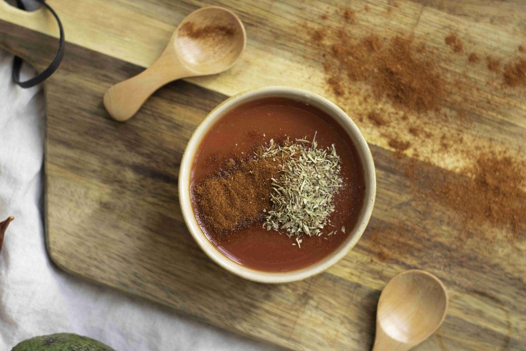 A bowl of tomato soup topped with dried herbs and ground spices sits on a wooden cutting board, next to two wooden spoons and a dusting of spice—perfect alongside the best beans and rice or your favorite vegetarian enchiladas.