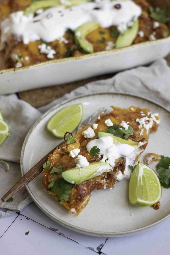 A plate with a serving of the best vegetarian enchiladas casserole, topped with avocado slices, sour cream, cheese, and cilantro is shown. Lime wedges and a fork are on the plate, with the casserole dish visible in the background.