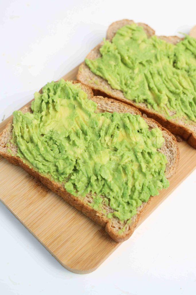 Two slices of whole grain bread topped with mashed avocado and a sprinkle of queso fresco create a delicious Mexican Avocado Toast, served on a light wooden cutting board against a white background.