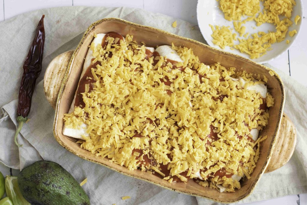 A baking dish filled with vegetarian enchiladas covered in shredded cheese, ready to be baked. The dish is placed on a cloth with an avocado, a dried chili, lime wedges, and a bowl of extra shredded cheese nearby.