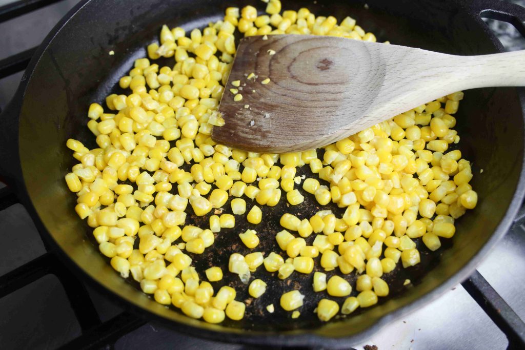 A wooden spatula stirs yellow corn kernels in a black skillet on a stovetop, perfect for topping Mexican Avocado Toast or pairing with crumbled Queso Fresco.