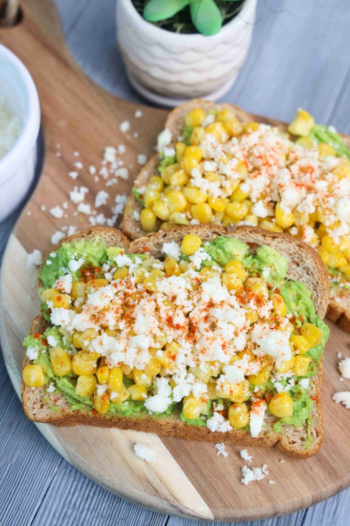 Two slices of whole grain toast topped with mashed avocado, corn kernels, and crumbled queso fresco create a vibrant Mexican Avocado Toast. A small plant and a bowl are partially visible in the background on the wooden board.