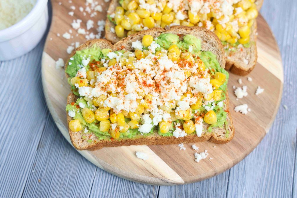 Two slices of Mexican Avocado Toast are topped with smashed avocado, sweet corn, and crumbled queso fresco, finished with a sprinkle of red seasoning and scattered cheese crumbs on a wooden board.