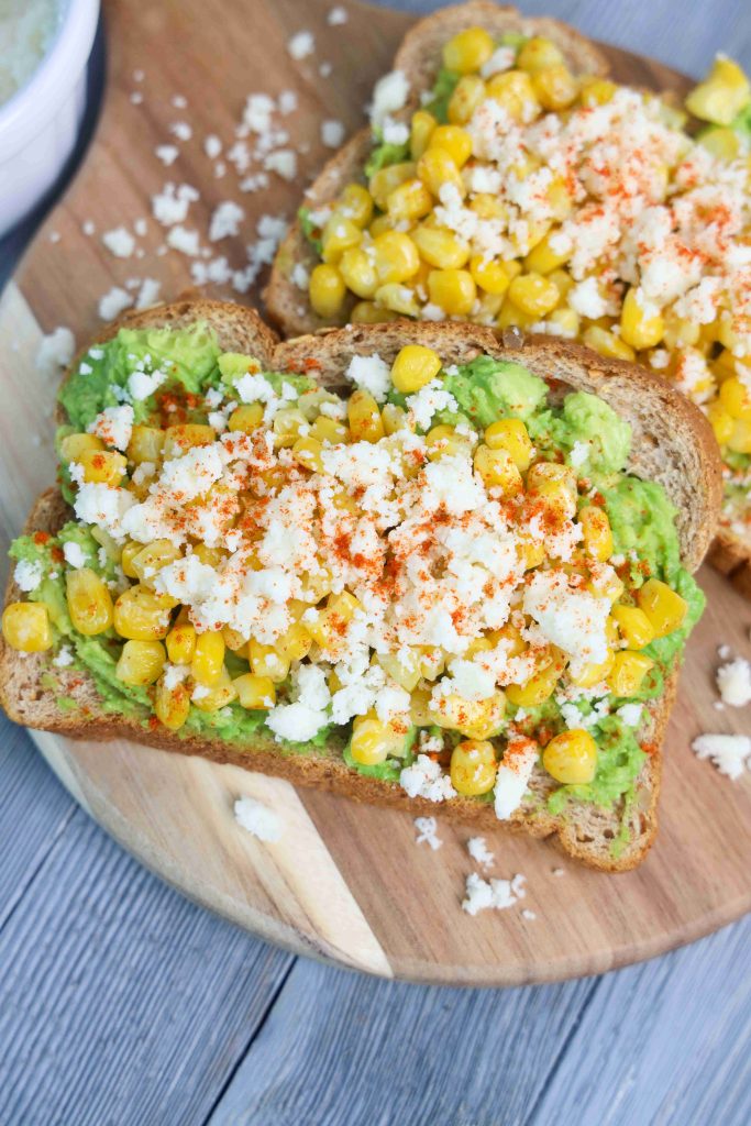 Enjoy Mexican Avocado Toast: two slices of whole grain toast topped with mashed avocado, roasted corn, crumbled queso fresco, and a sprinkle of red chili powder, served on a wooden board.
