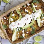 A casserole dish filled with the best enchiladas topped with sliced avocado, sour cream, crumbled cheese, and cilantro. Lime wedges and half an avocado sit nearby on a light-colored table. Perfect alongside beans and rice.