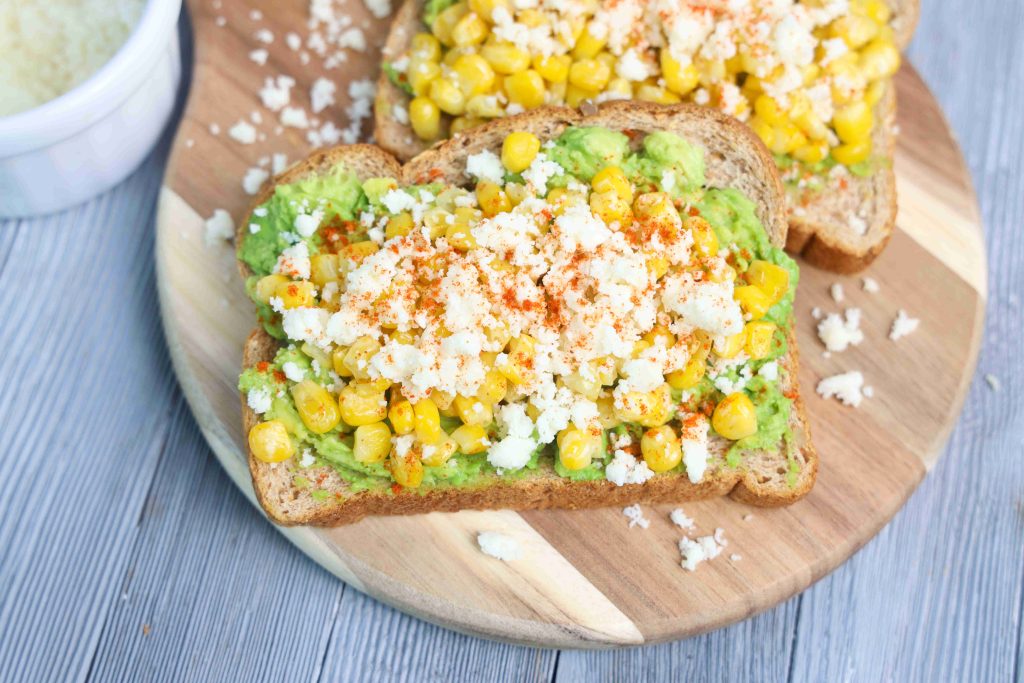 Two slices of Mexican avocado toast topped with mashed avocado, corn, crumbled queso fresco, and a sprinkle of chili powder sit on a round wooden board. A small white bowl with extra cheese is nearby.