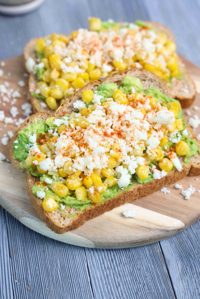 Two slices of Mexican Avocado Toast topped with mashed avocado, sweet corn kernels, crumbled queso fresco, and a sprinkle of paprika, served on a wooden board.