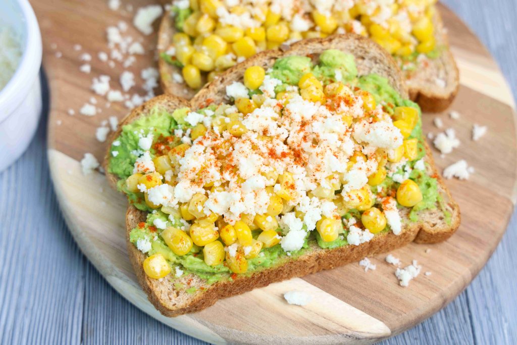 Two slices of Mexican avocado toast topped with mashed avocado, corn, crumbled queso fresco, and a sprinkle of chili powder, served on a wooden board. Some crumbs and corn kernels are scattered around.