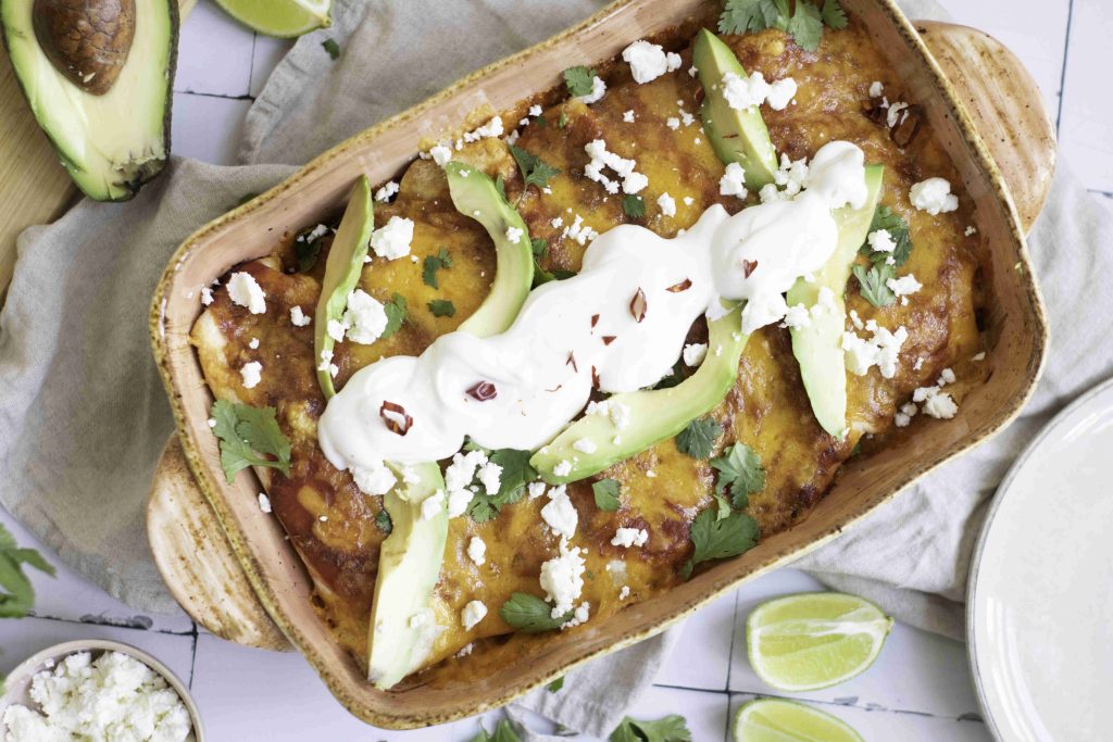 A baking dish filled with vegetarian enchiladas topped with melted cheese, sliced avocado, sour cream, fresh cilantro, and crumbled cheese. Lime wedges and an avocado are nearby on a white tile surface.