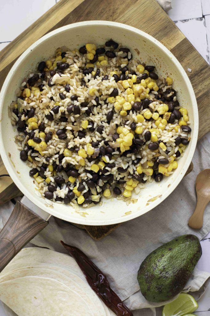 A skillet contains a mixture of black beans and rice, perfect for making the best vegetarian enchiladas. Surrounding it are tortillas, a wooden spoon, an avocado, lime wedges, and dried chilies on a wooden board and cloth.