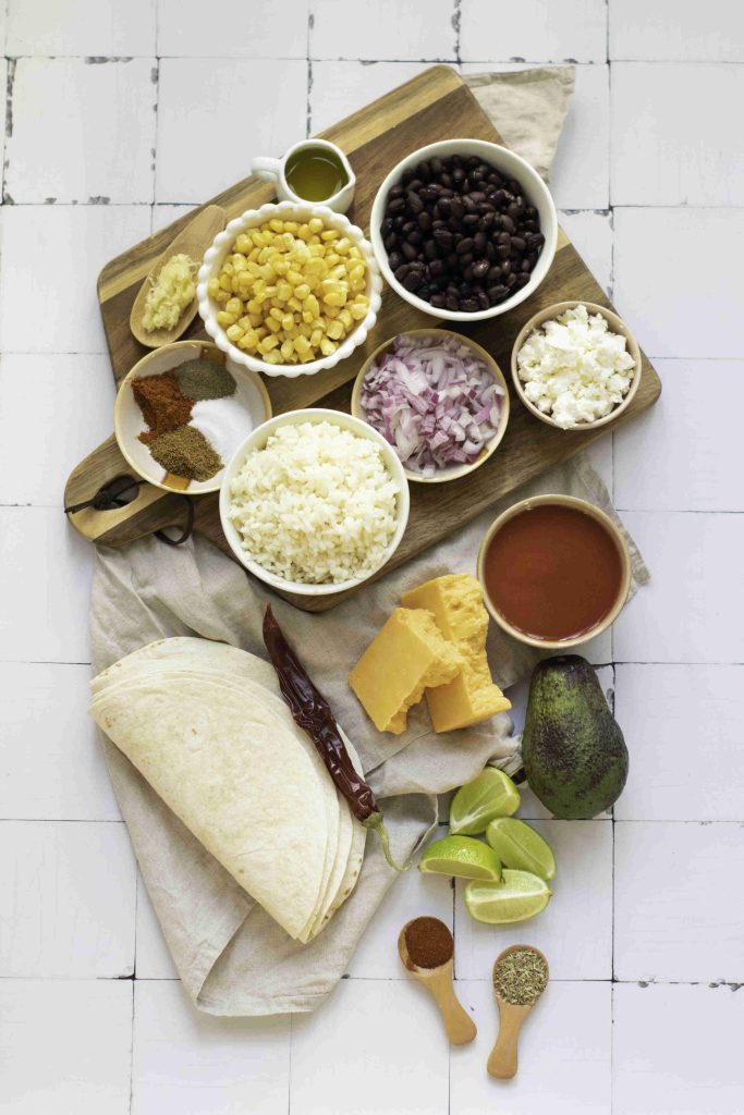 A top-down view of burrito ingredients on a wooden board, including tortillas, corn, black beans and rice, onions, cheese, avocado, lime wedges, red sauce, and various spices—perfect for making the best vegetarian enchiladas.