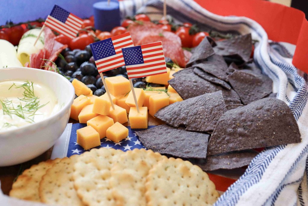 A festive 4th of July Charcuterie Board with crackers, cheddar cubes, blue corn chips, berries, and small American flags on a blue-and-white striped cloth. A bowl of dip and salami slices complete this patriotic spread.