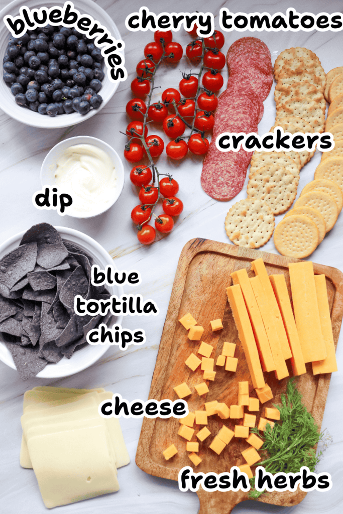 A flat lay of a 4th of July Charcuterie Board with labeled ingredients: a bowl of blueberries, cherry tomatoes on the vine, two types of crackers, dip, blue tortilla chips, sliced and cubed cheese, fresh herbs, and deli cheese.
