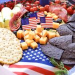 A festive 4th of July Charcuterie Board featuring crackers, cheese cubes, blue corn chips, rolled cheese, deli meats, cherry tomatoes, and small American flags on a star-spangled cloth.
