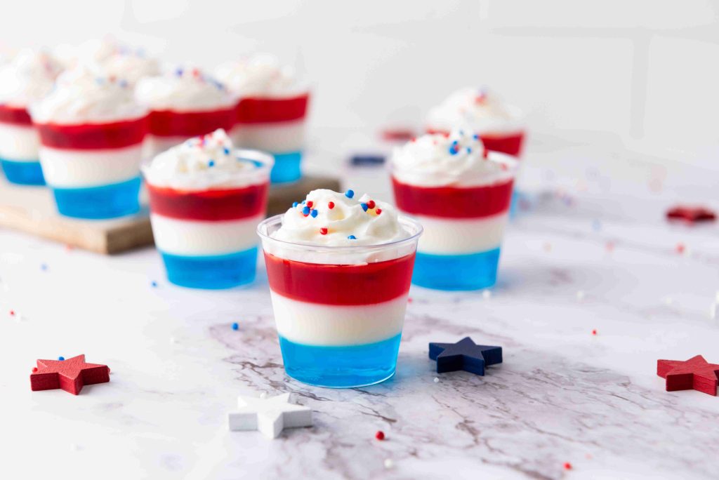Individual cups filled with layered blue, white, and red Vodka Jello Shots, topped with whipped cream and festive sprinkles. Decorative stars are placed on a marble surface around the cups—perfect for your 4th of July celebration.