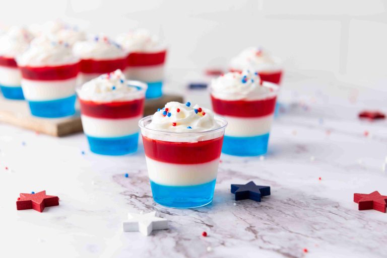Individual cups filled with layered blue, white, and red Vodka Jello Shots, topped with whipped cream and festive sprinkles. Decorative stars are placed on a marble surface around the cups—perfect for your 4th of July celebration.