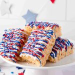 Rice Krispies treats stacked on a white cake stand, decorated with red, white, and blue icing plus festive star-shaped sprinkles—perfect 4th of July treats. Patriotic-themed star decorations hang in the background.
