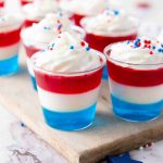 Small cups filled with layered red, white, and blue 4th of July Jello Shots, topped with whipped cream and festive sprinkles, arranged on a wooden board.