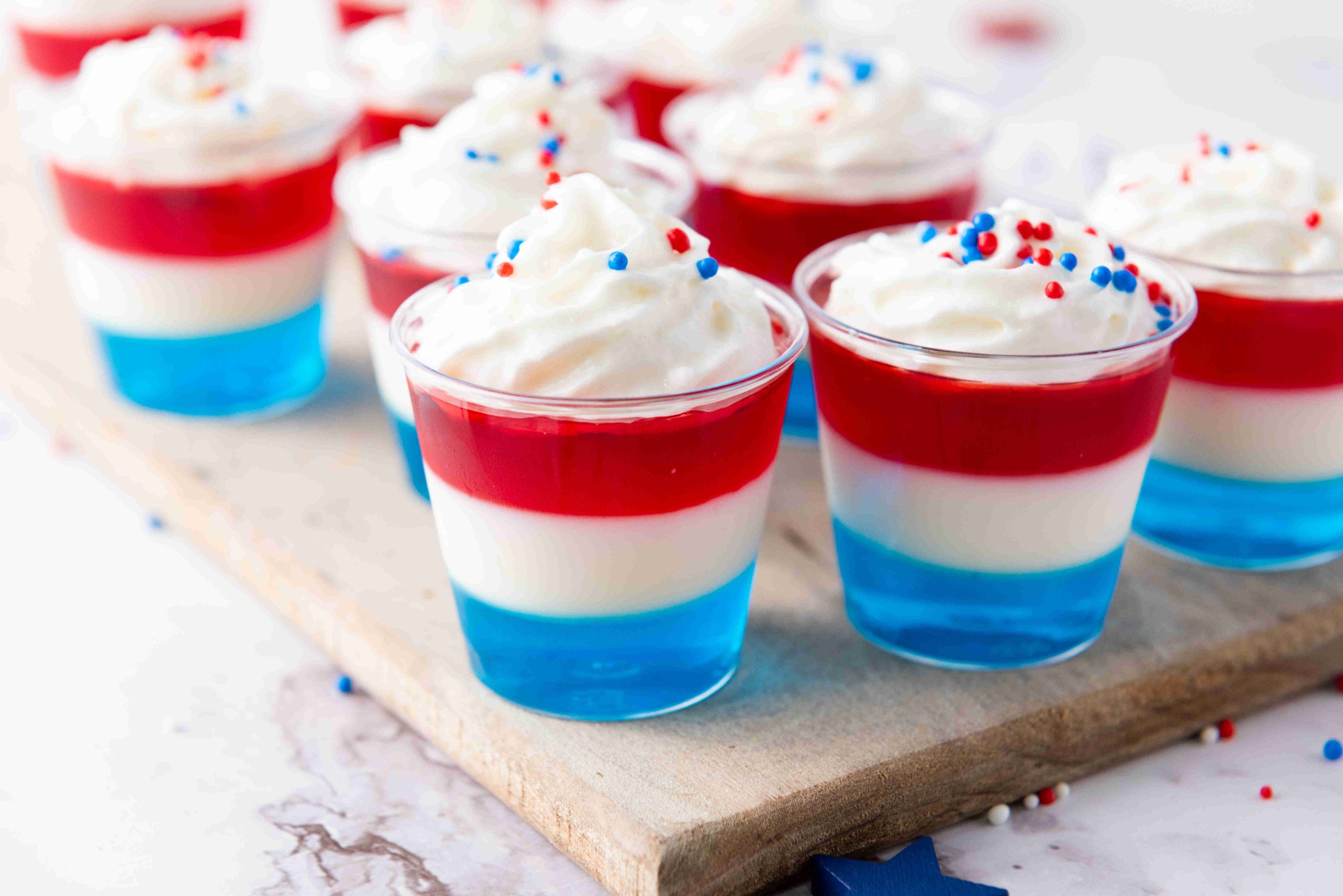 Small cups filled with layered red, white, and blue 4th of July Jello Shots, topped with whipped cream and festive sprinkles, arranged on a wooden board.