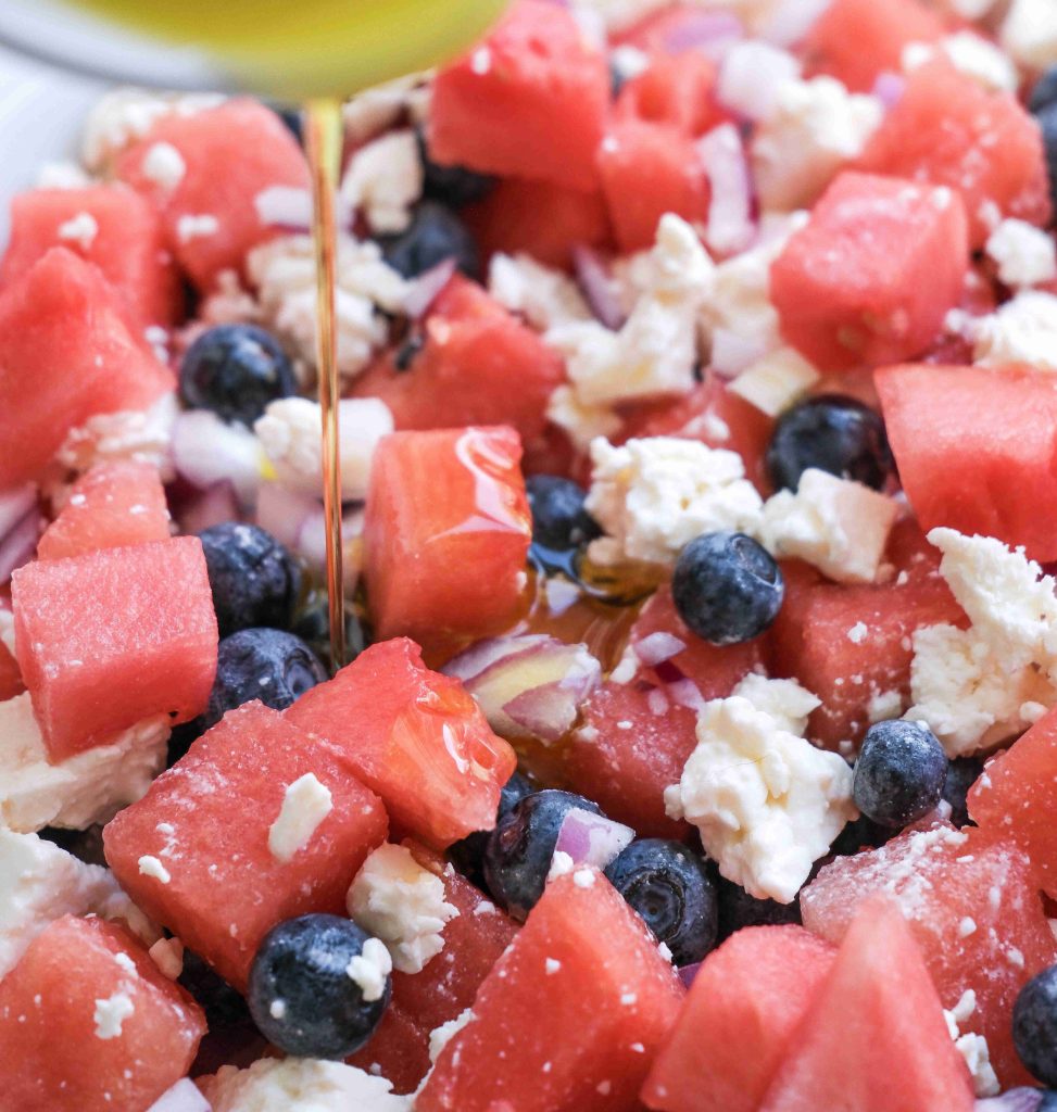 A close-up of a Red White Blue Salad made with cubes of watermelon, blueberries, crumbled feta cheese, and finely chopped onions, with a drizzle of dressing being poured over the top—perfect for your next Summer BBQ.