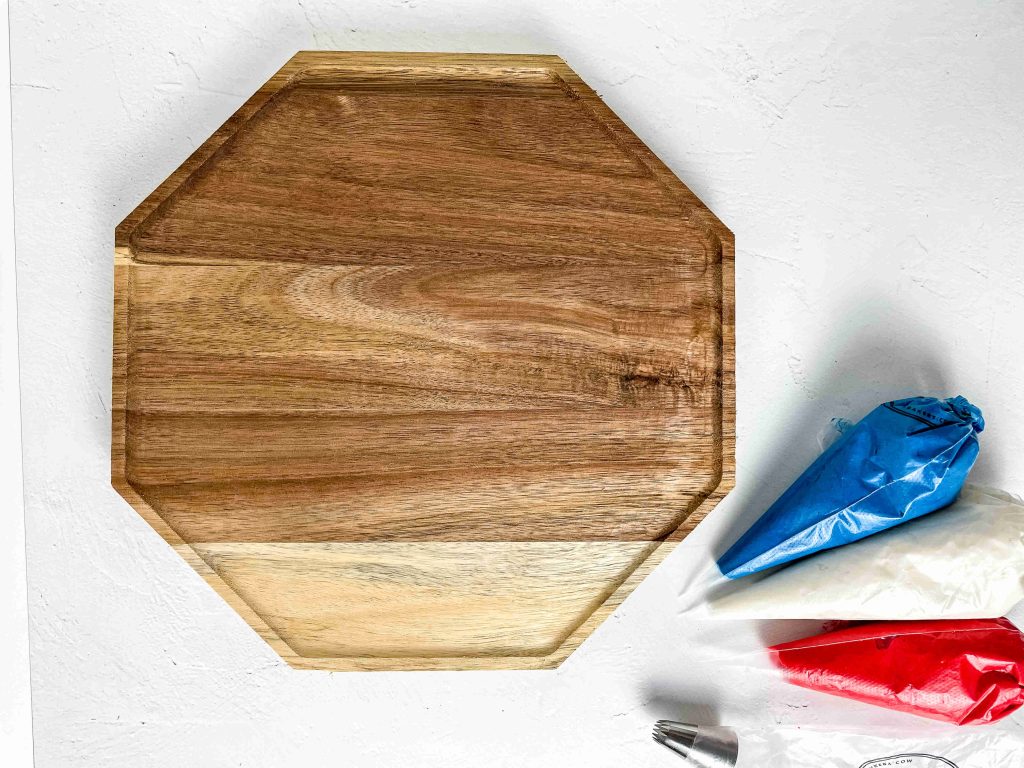 A wooden octagonal charcuterie board on a white surface, with piping bags filled with red, white, and blue icing—perfect for creating festive 4th of July treats—plus a metal icing tip placed to the right of the board.