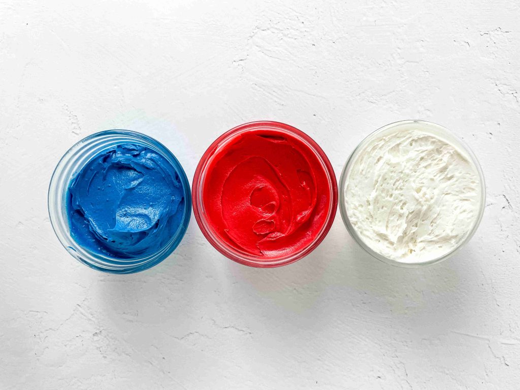 Three glass bowls filled with blue, red, and white frosting are arranged in a row on a white textured surface—perfect for adding color to your 4th of July charcuterie board.