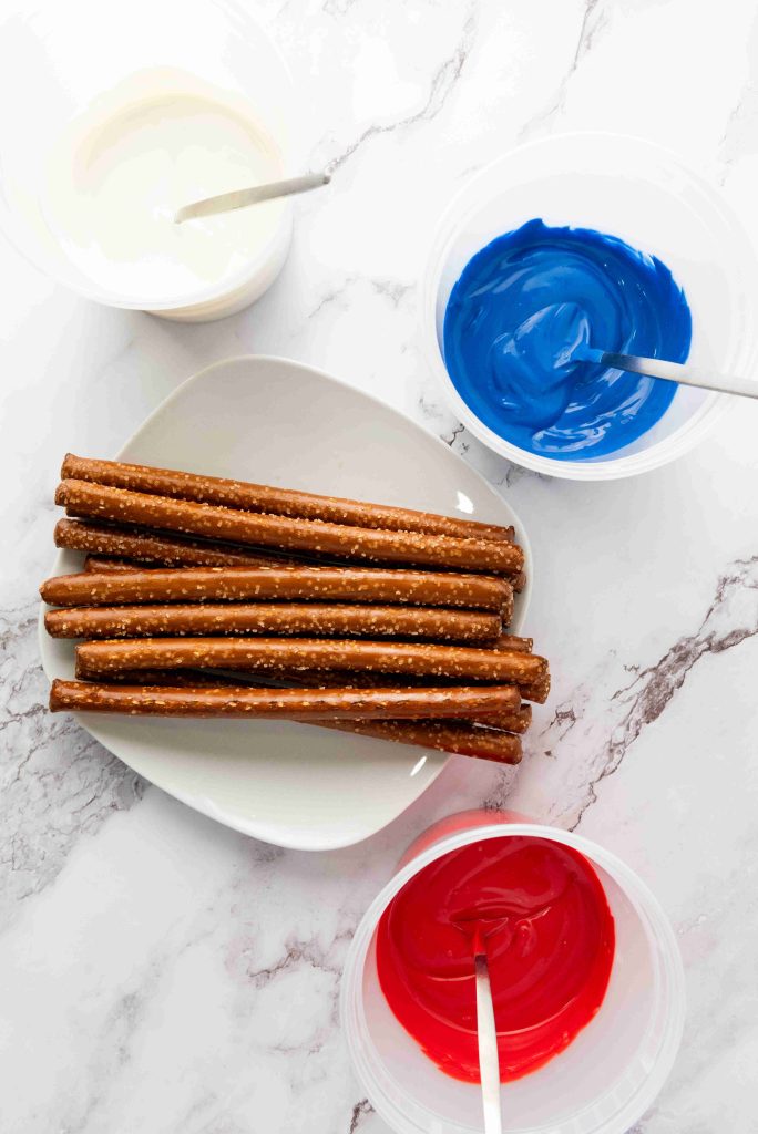 A plate of Patriotic Pretzel Rods is surrounded by three bowls of melted white, blue, and red chocolate, each with a spoon for dipping—a festive 4th of July treat on a marble surface.